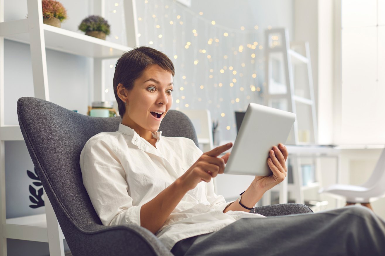 Overjoyed Adult Woman Reading Exciting News on Tablet Sitting in Comfortable Armchair at Home